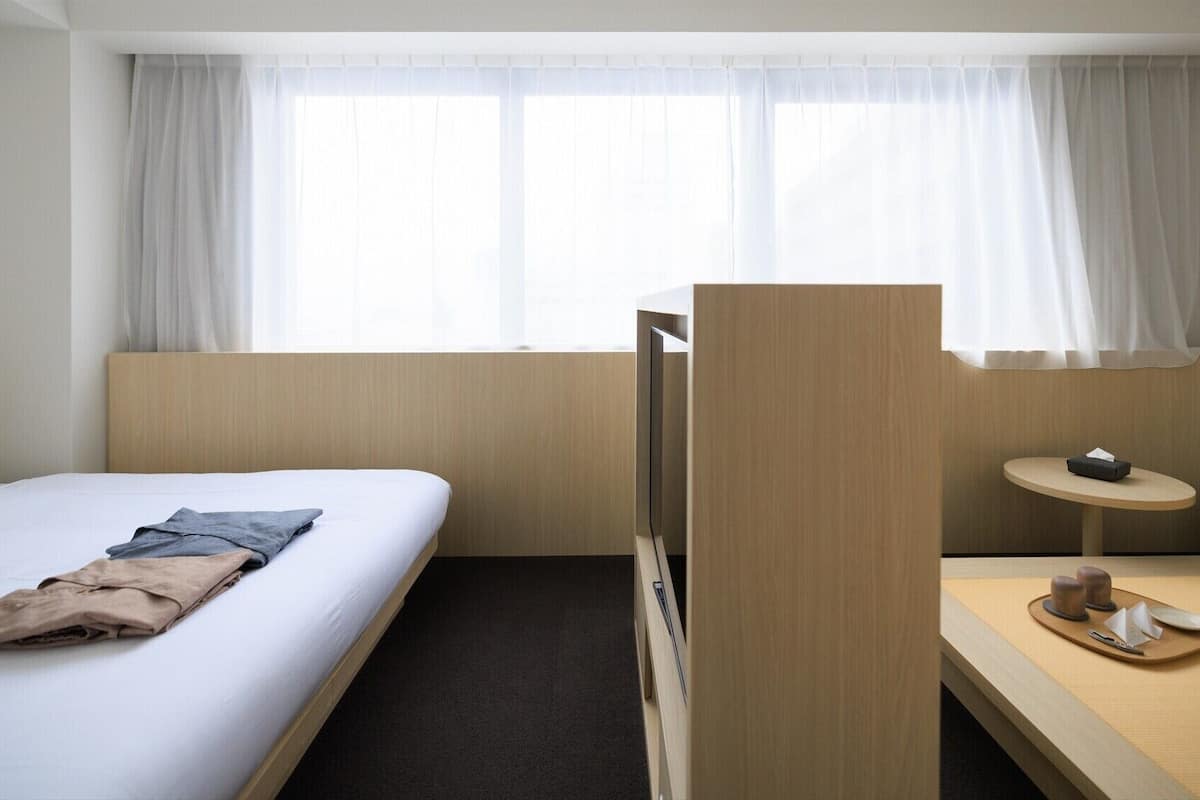 Una habitación de hotel con cama, escritorio y bandeja de objetos, y ventana con cortinas.