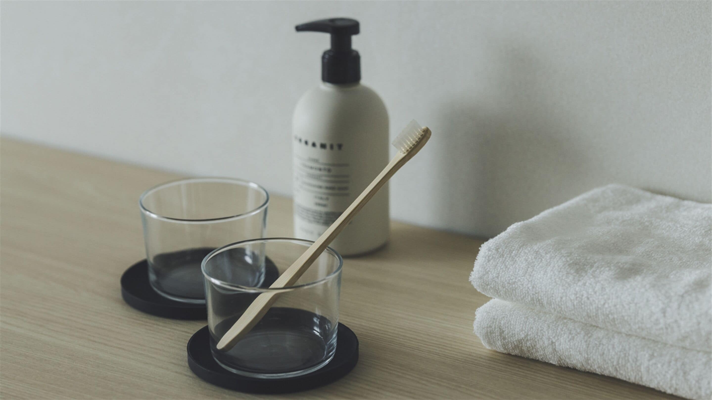 A bathroom with a wooden floor, two clear glasses on black coasters, a white towel, and a bottle of shampoo.