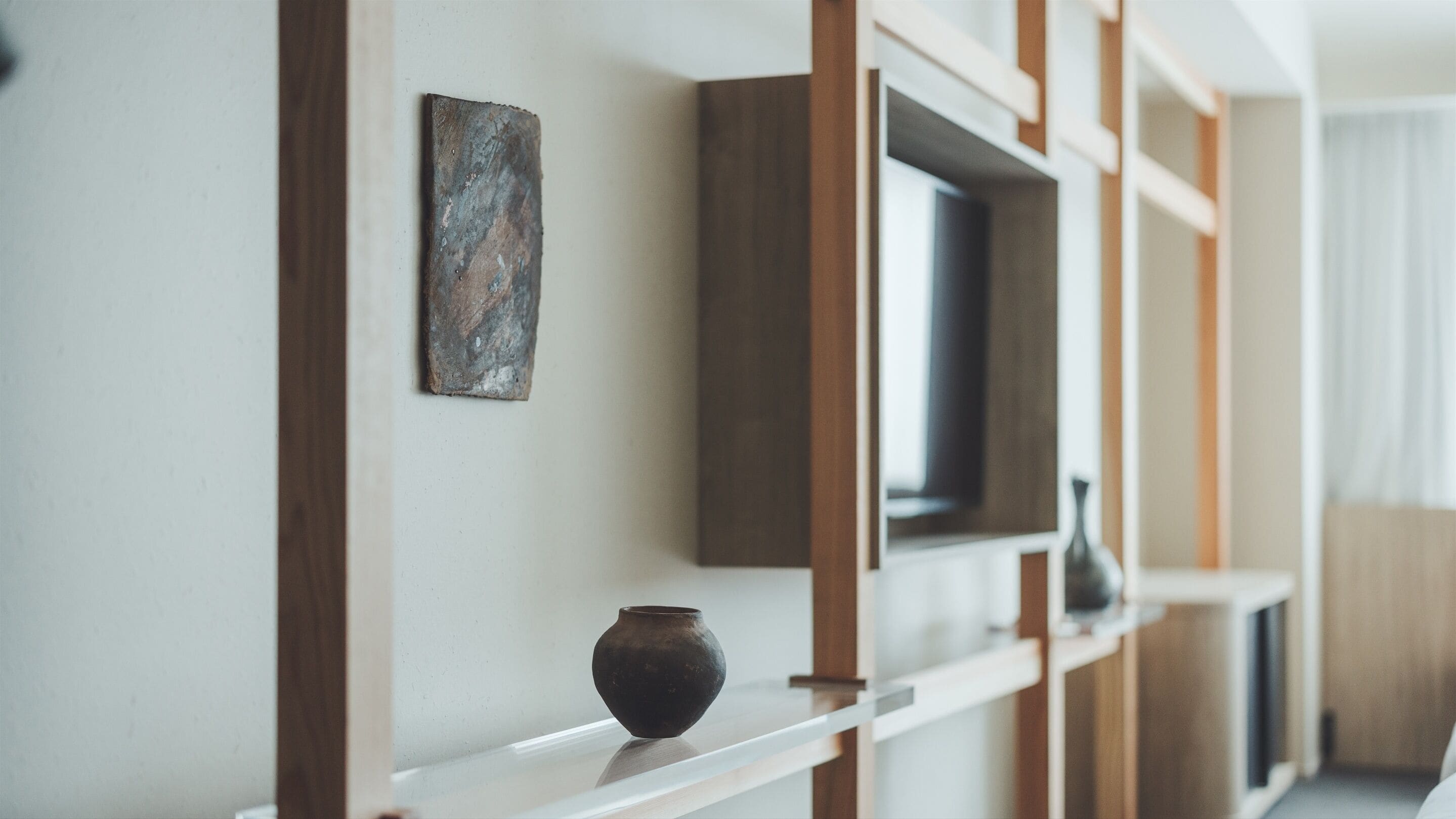 A modern interior with a minimalist design, featuring a wooden shelf with a vase and a painting, and a television mounted on the wall.