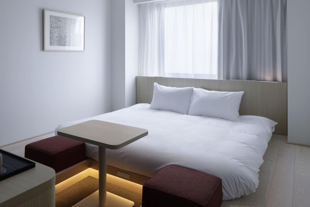 Una habitación de hotel moderna con una cama bien hecha, una mesita de noche pequeña con una lámpara y una ventana con cortinas traslúcidas.