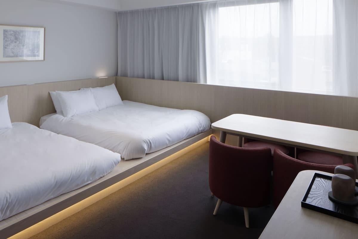 Habitación de hotel con dos camas individuales, un escritorio, una silla y un ventanal con cortinas.