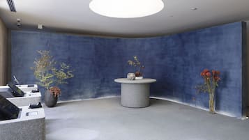 Ein Empfangsbereich mit einer blauen, bogenförmigen Wand, einem runden Tisch in der Mitte mit einer Vase und einer Sitzecke mit einer Bank und einem kleinen Tisch.