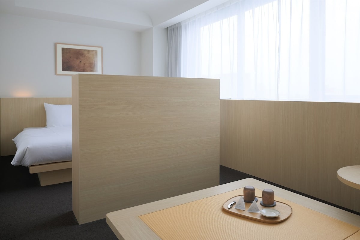 Habitación de hotel con cama, una mesita con bandeja, mesita de noche y ventana con cortinas.