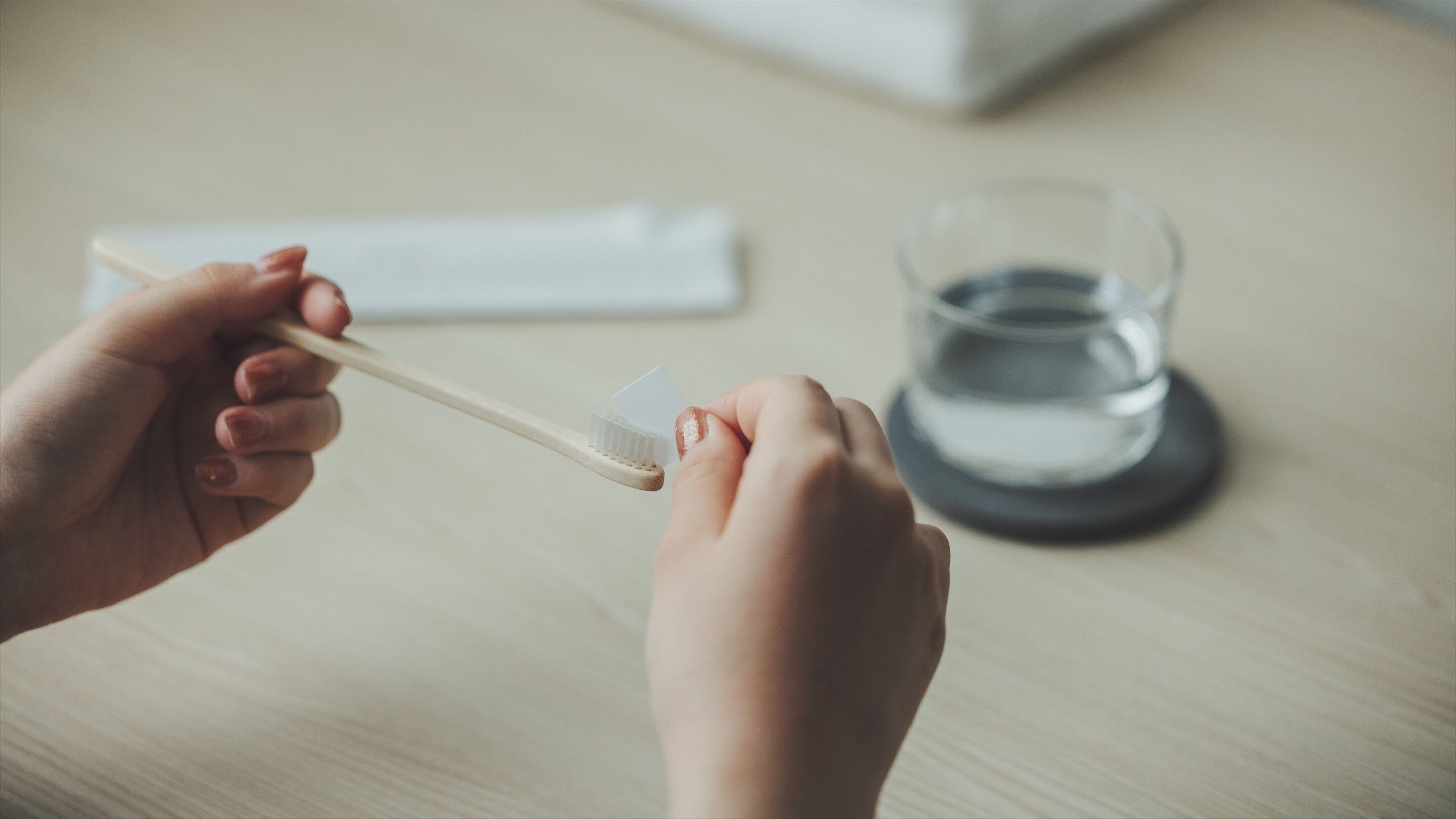 Une main tenant une brosse à dents et un morceau de papier, et un verre d’eau posé sur une table.
