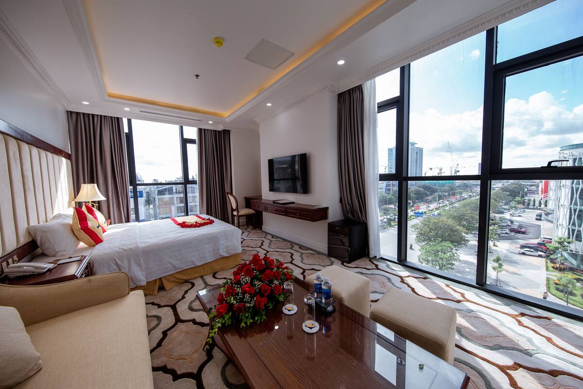 Ein Hotelzimmer mit einem großen Bett, einem Schreibtisch mit Blumen in einer Vase, einem Fernseher an der Wand und Blick auf die Stadt durch große Fenster.
