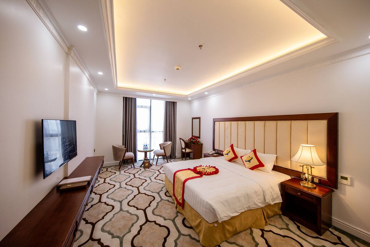 Ein Hotelzimmer mit einem großen Bett, Nachttisch, Lampe, Fernseher und einem Schreibtisch mit Stühlen.