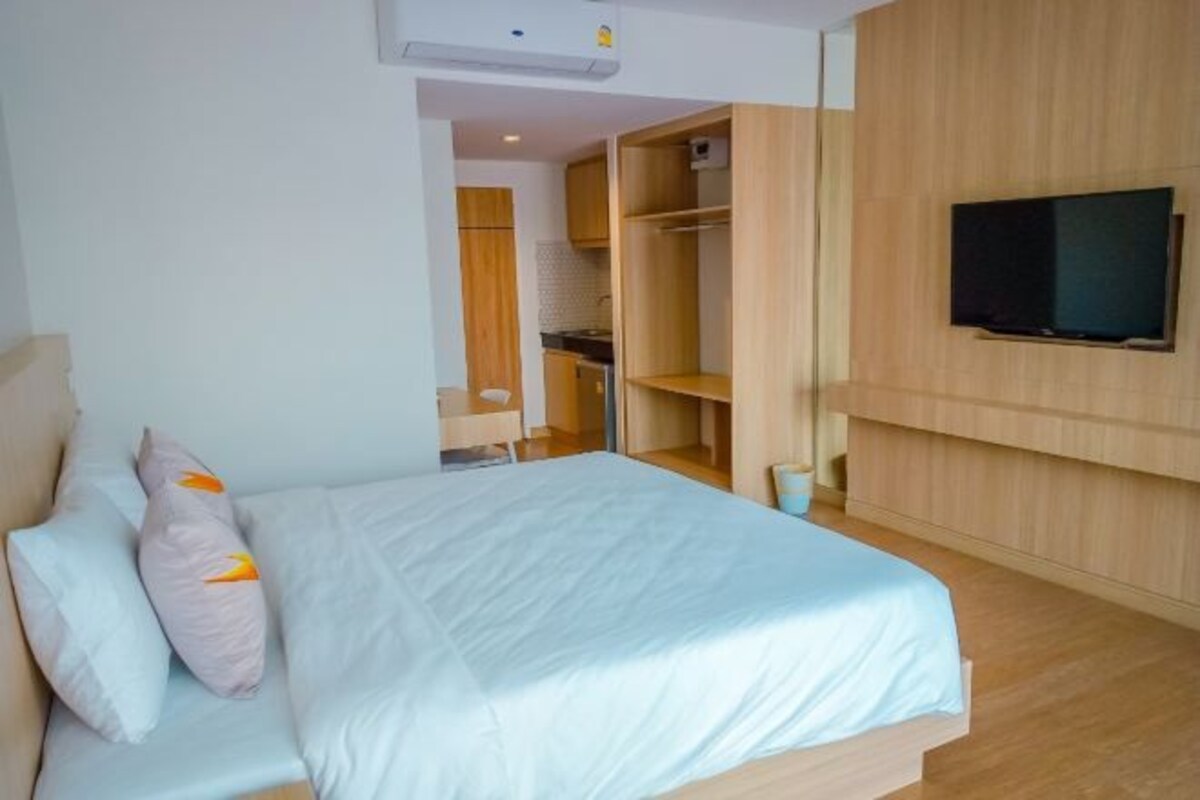 A bedroom with a bed, a TV mounted on the wall, and a kitchenette in the background.