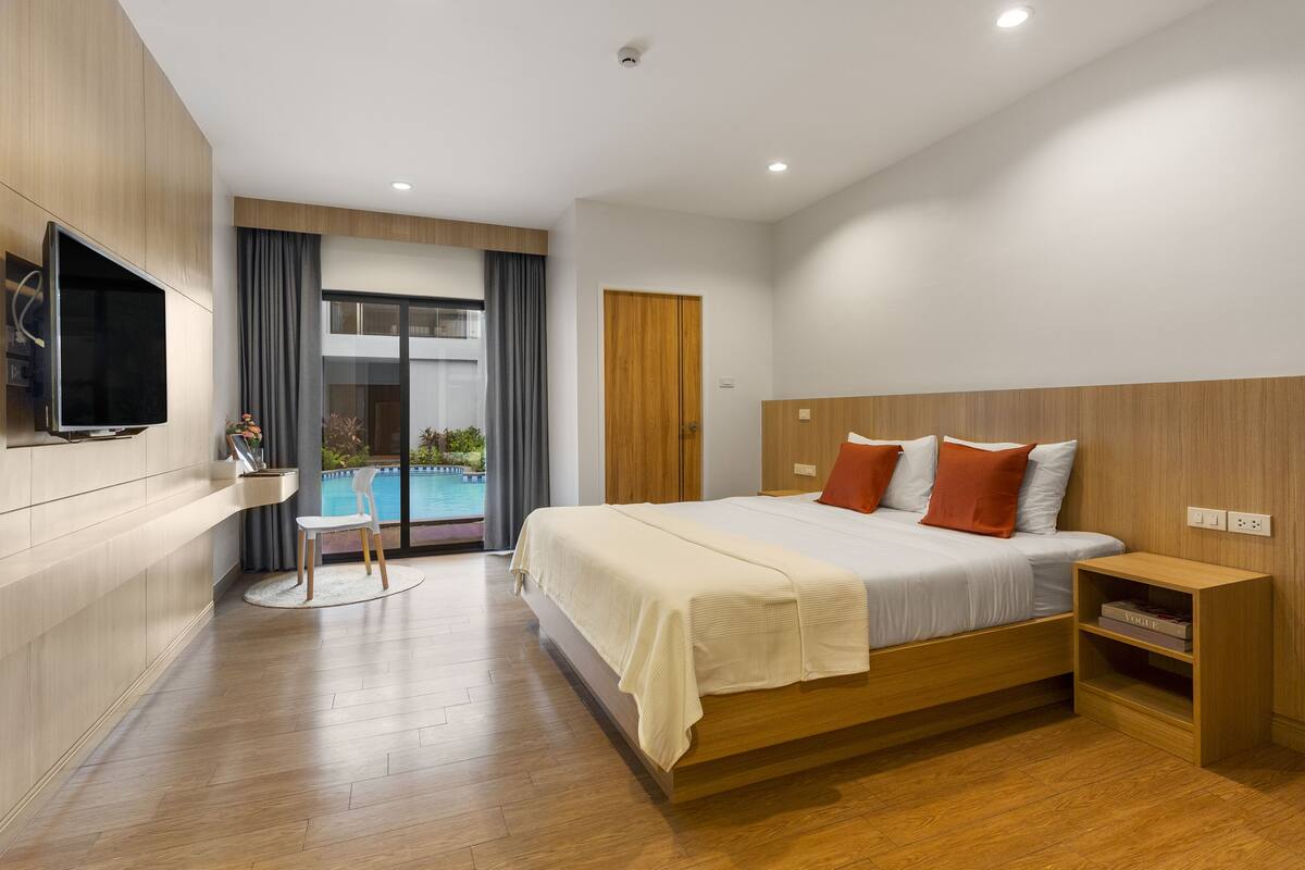A modern hotel room with a large bed, a TV mounted on the wall, a wooden nightstand, and a view of a swimming pool.