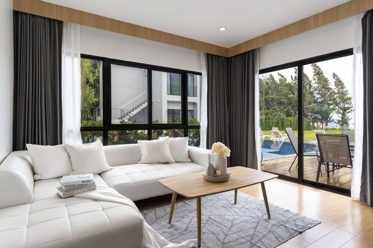 A modern living room with a white sofa, a coffee table, and a view of an outdoor pool area through large windows.