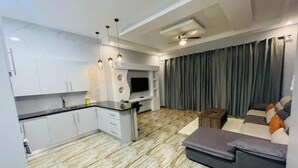 A modern kitchen with white cabinets, a black countertop, and a built-in TV. There is a grey sofa with cushions and a ceiling fan above.