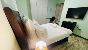 A neatly made bed with a headboard, a bedside lamp, a TV mounted on the wall, and a wardrobe in the background. - Ibex Meanwood suite (Lusaka)