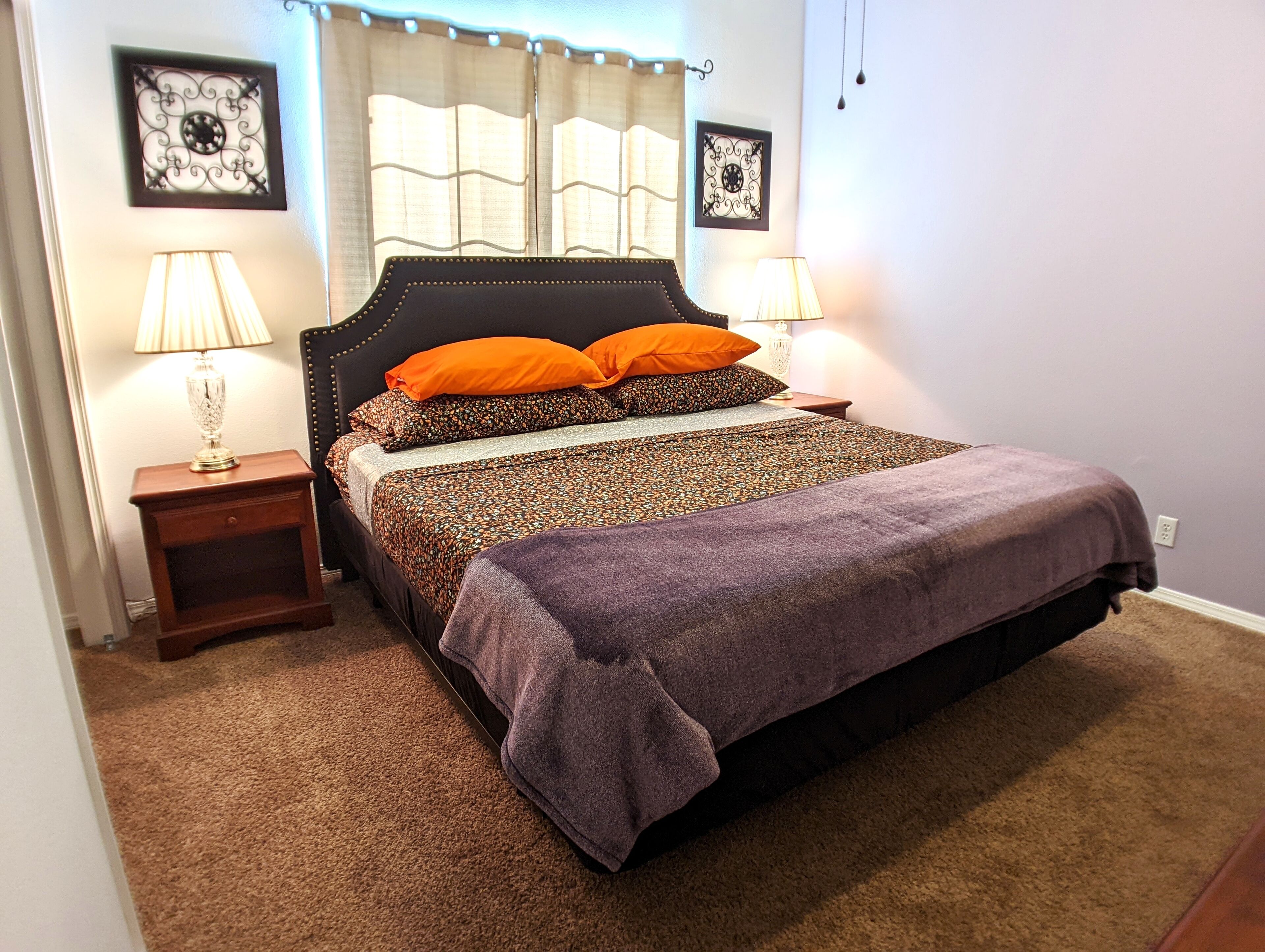 Cama bem arrumada com colcha estampada e duas almofadas laranjas, acompanhada por duas mesas de cabeceira com abajures, em um quarto com piso carpetado e decoração nas paredes.
