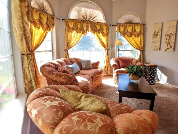 A living room with a sofa set, a coffee table, and a lamp. There are curtains and a window with a view.