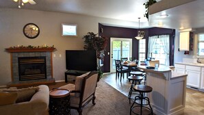 Sala de estar con chimenea, televisor y zona de estar. Se observa un comedor con una cocina al fondo.