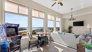 A spacious living room with a large window, a ceiling fan, a sofa, a TV, and a gaming console.