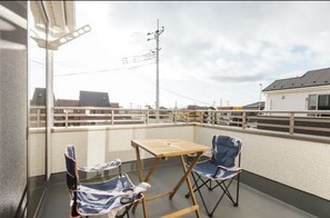 Een balkon met een tafel en twee stoelen, een witte muur en een uitzicht op gebouwen en hoogspanningslijnen.