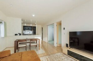 A modern living room with a large flat-screen TV, a dining area with a table and chairs, and a kitchen in the background.