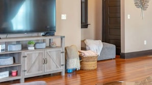 A living room with a flat-screen TV on a wooden stand, a grey couch, a wicker basket, and a decorative leaf wall piece. - Remodeled historic family home w/ full kitchen & garage parking (St. Louis)