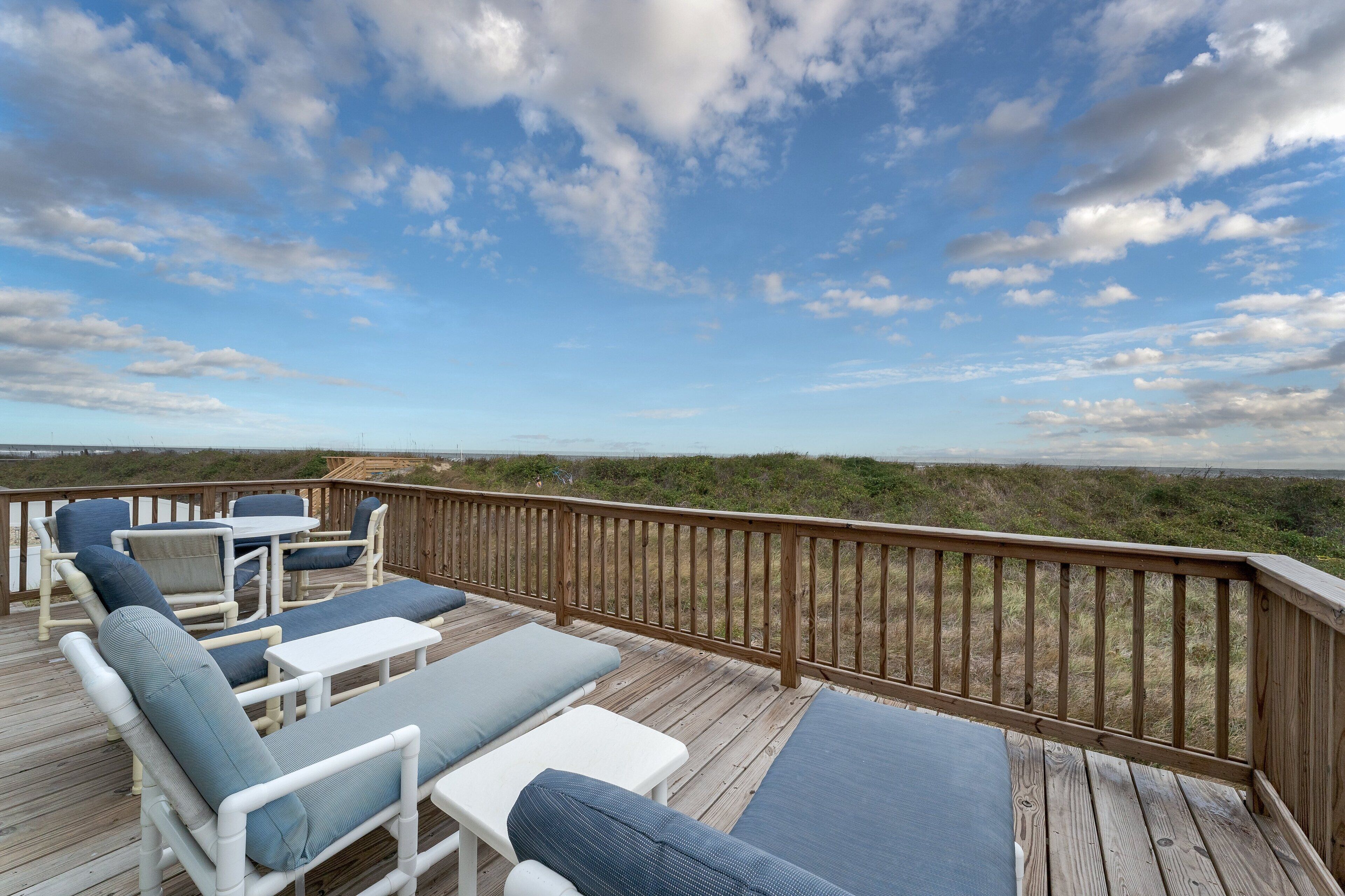 A wooden deck with outdoor furniture, including lounge chairs and a table, overlooking a beach and dunes.