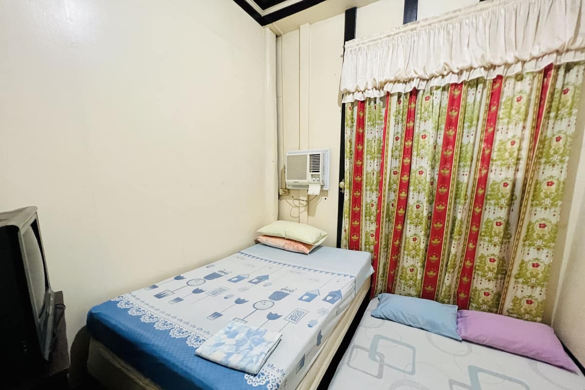 Una habitación pequeña y sencilla con dos camas, aire acondicionado instalado en la pared y una ventana con cortinas de flores.