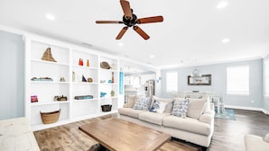 A spacious living room with a wooden coffee table, a beige sofa, and a white bookshelf filled with decorative items.