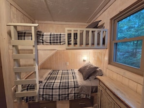 A bunk bed with a ladder, a single bed with a plaid blanket, a nightstand, and a window with a forest view.
