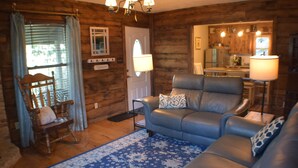 A cozy living room with a blue patterned rug, a leather sofa, a wooden rocking chair, and a kitchen visible through an open door. - The Linville Community, a restful retreat w/ convenient access to the mountains (Newland)