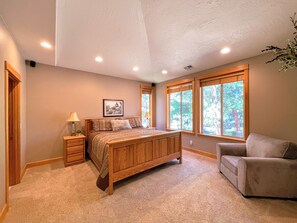 A bedroom with a wooden bed, a nightstand, a chair, and large windows with a view of trees. - ★ Wonderful 5bdrm Getaway -Chef's kitchen -Minutes to river + AC +SHARC passes ★ (Sunriver)