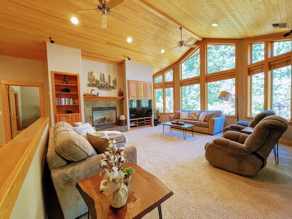 A spacious living room with a fireplace, large windows, and a cozy seating area. - ★ Wonderful 5bdrm Getaway -Chef's kitchen -Minutes to river + AC +SHARC passes ★ (Sunriver)