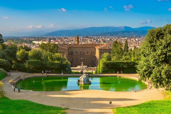 A historic building with a fountain, surrounded by greenery and a view of a cityscape.