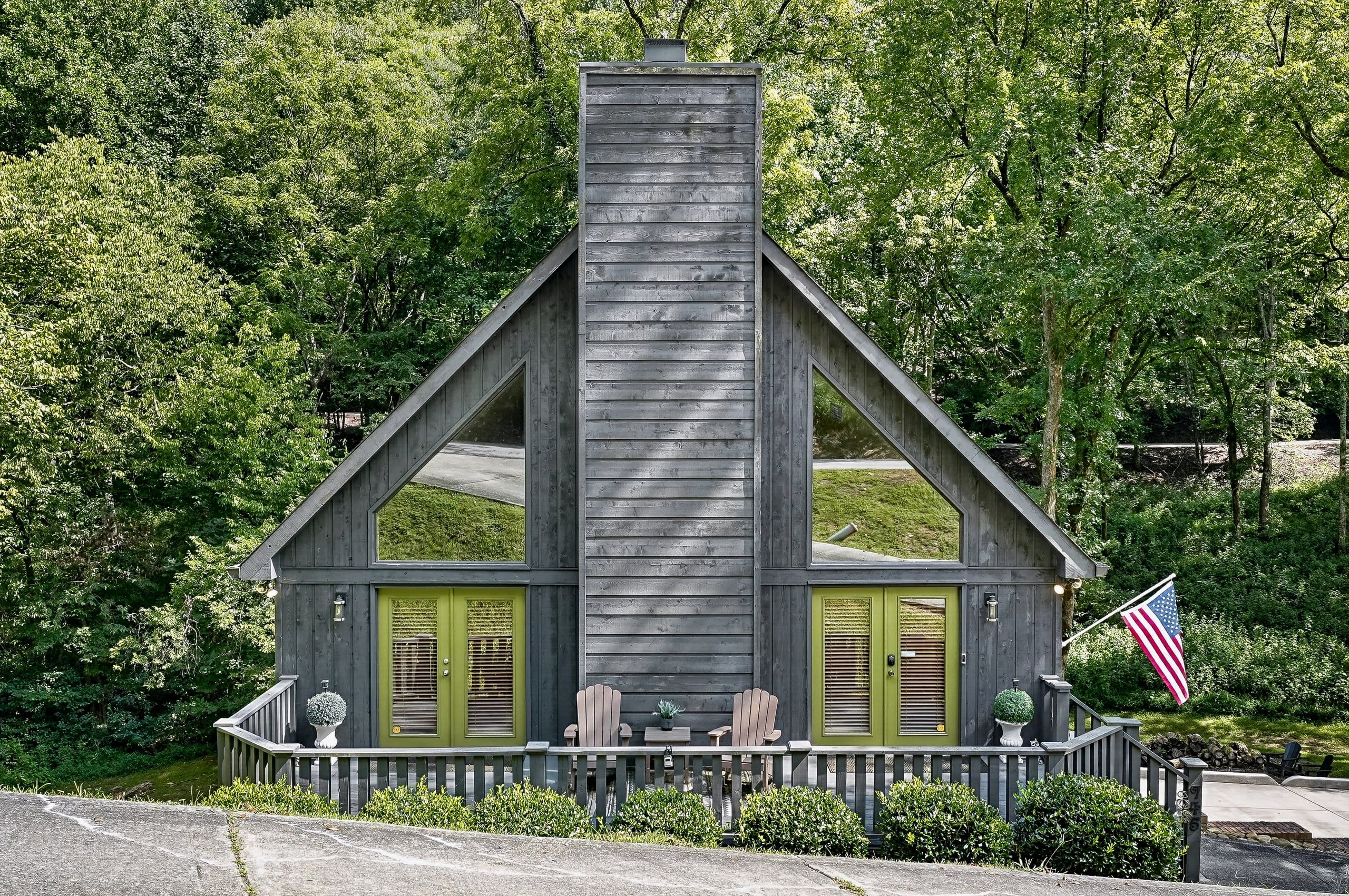 A wooden cabin with a chimney, two green doors, and a porch with Adirondack chairs.