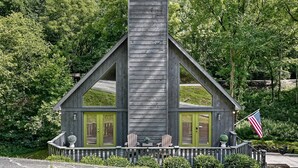Uma cabana de madeira com chaminé, duas portas verdes e um alpendre com cadeiras Adirondack.