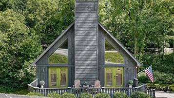 A wooden cabin with a chimney, two green doors, and a porch with Adirondack chairs.