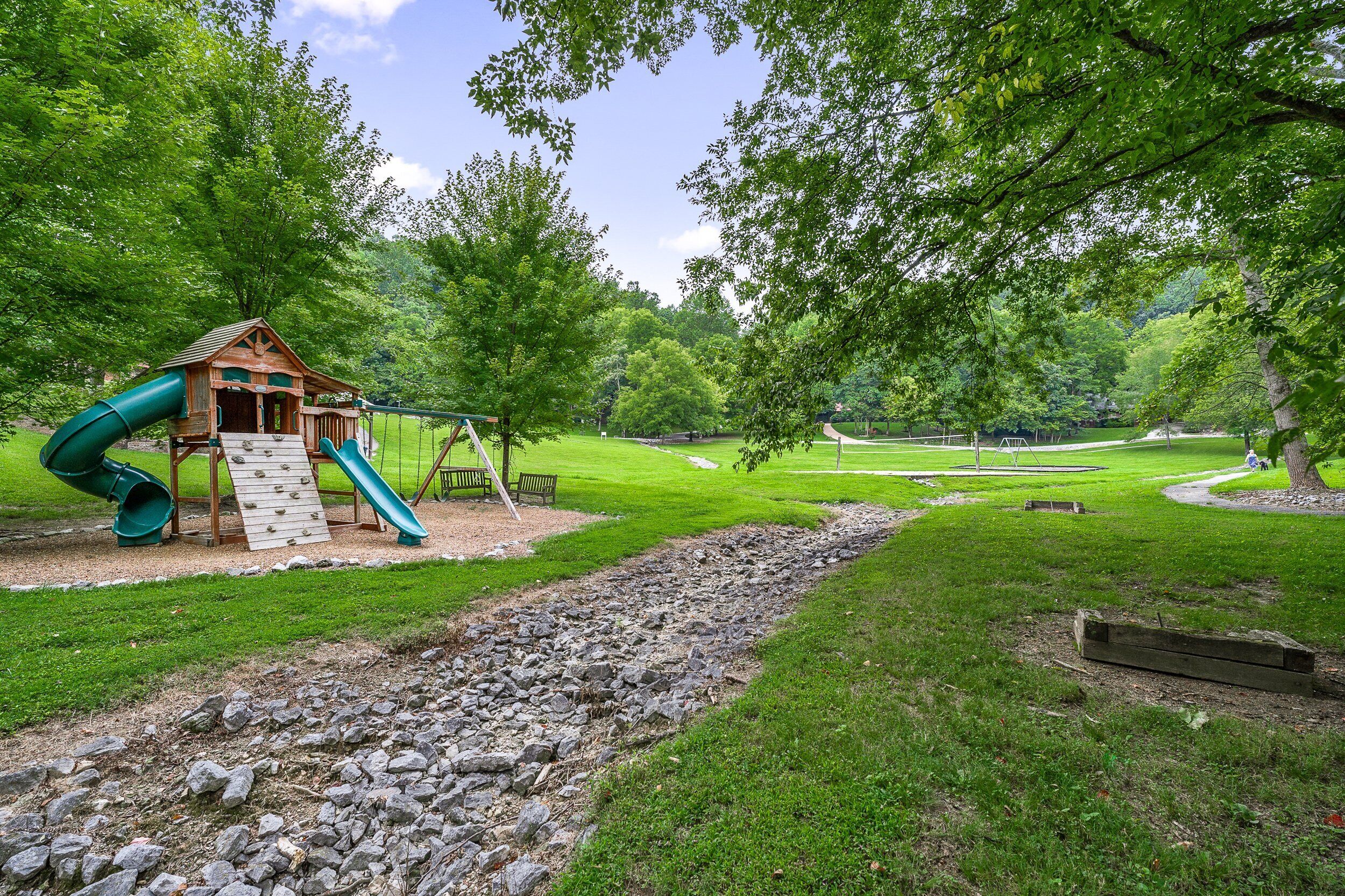 A playground with a wooden playhouse, slides, and swings surrounded by lush green grass and trees.
