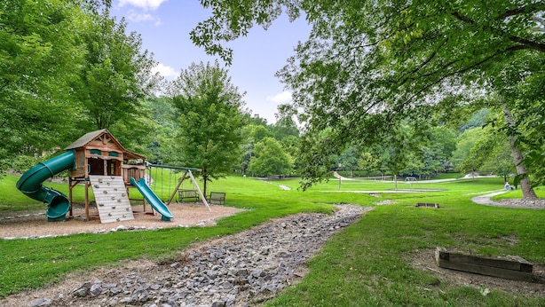 A playground with a wooden playhouse, slides, and swings surrounded by lush green grass and trees.