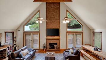 A spacious loft with a high ceiling, exposed wooden beams, a brick fireplace, and large windows offering views of greenery.