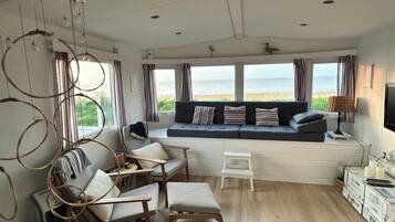 Ein gemütliches Wohnzimmer mit großem Fenster mit Blick auf den Strand, einer Couch mit Streifenpolstern, Holzstühlen und einem kleinen Tisch.
