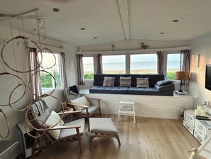 Ein gemütliches Wohnzimmer mit großem Fenster mit Blick auf den Strand, einer Couch mit Streifenpolstern, Holzstühlen und einem kleinen Tisch.