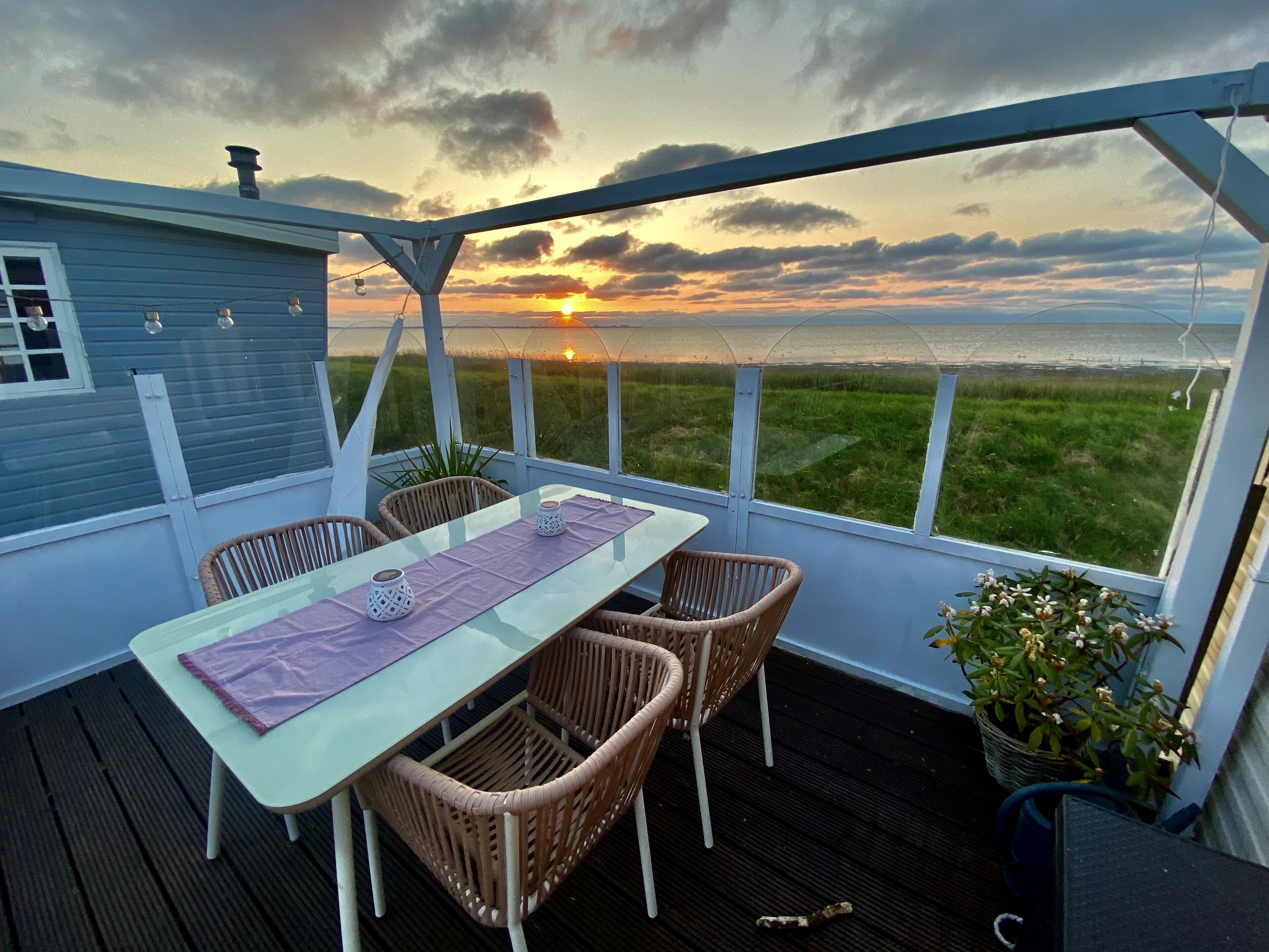 Un espace extérieur abrité, avec une table et des chaises, une vue sur le coucher de soleil sur l’eau et un champ d’herbe.