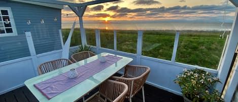 Un espace extérieur abrité, avec une table et des chaises, une vue sur le coucher de soleil sur l’eau et un champ d’herbe.