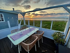 A covered outdoor area with a table and chairs, a view of the sunset over the water, and a grassy field.