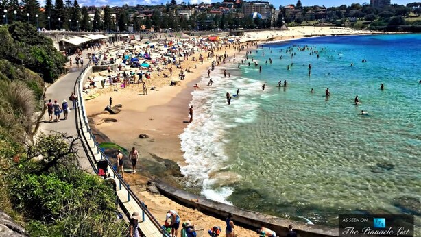 Une plage bondée, avec une eau d’un bleu limpide, un rivage sablonneux et une vue sur la ville en arrière-plan.