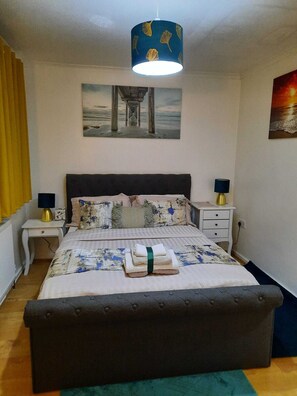 Quarto com cama, duas mesas de cabeceira, luminária pendente, pintura de um cais e janela com cortinas.