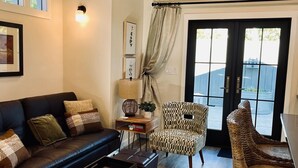 Una sala de estar con un sofá de cuero negro, un sillón estampado, una mesita auxiliar con una lámpara y una silla de mimbre.