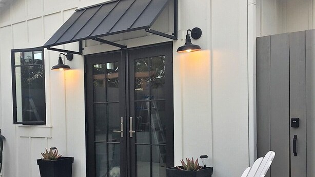 Un edificio blanco con toldo negro, dos puertas negras y un pequeño patio con una silla blanca y una mesa con una botella y vasos.
