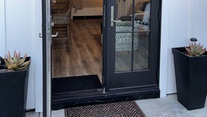 Una entrada moderna con puerta corredera de vidrio, un toldo negro y un felpudo. En el interior, hay una sala de estar con sofá, una mesa de centro y una alfombra.