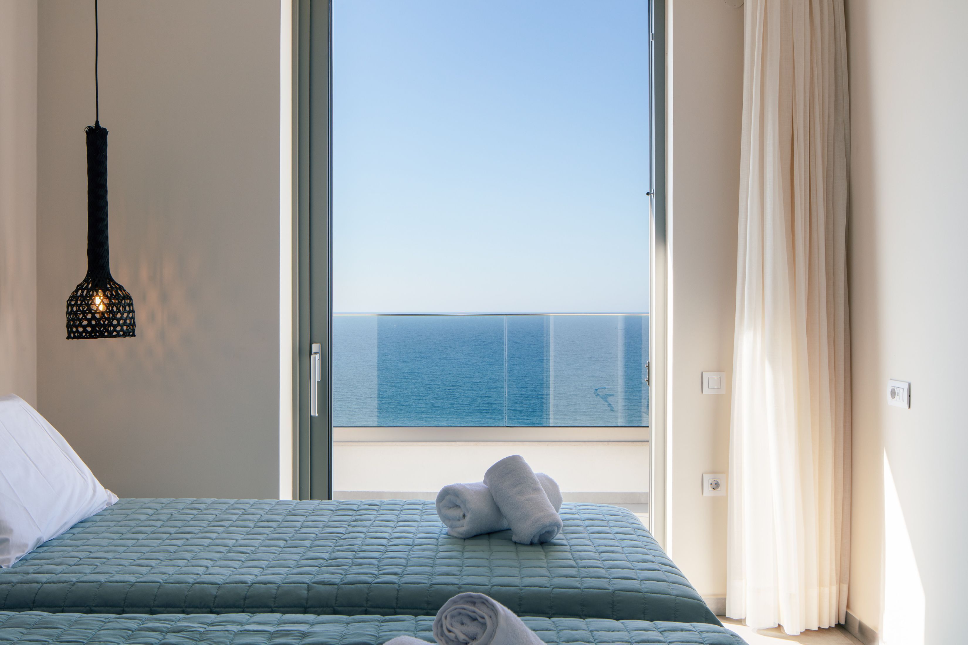 A hotel room with a bed, a hanging light fixture, a view of the sea through a window, and a curtain.