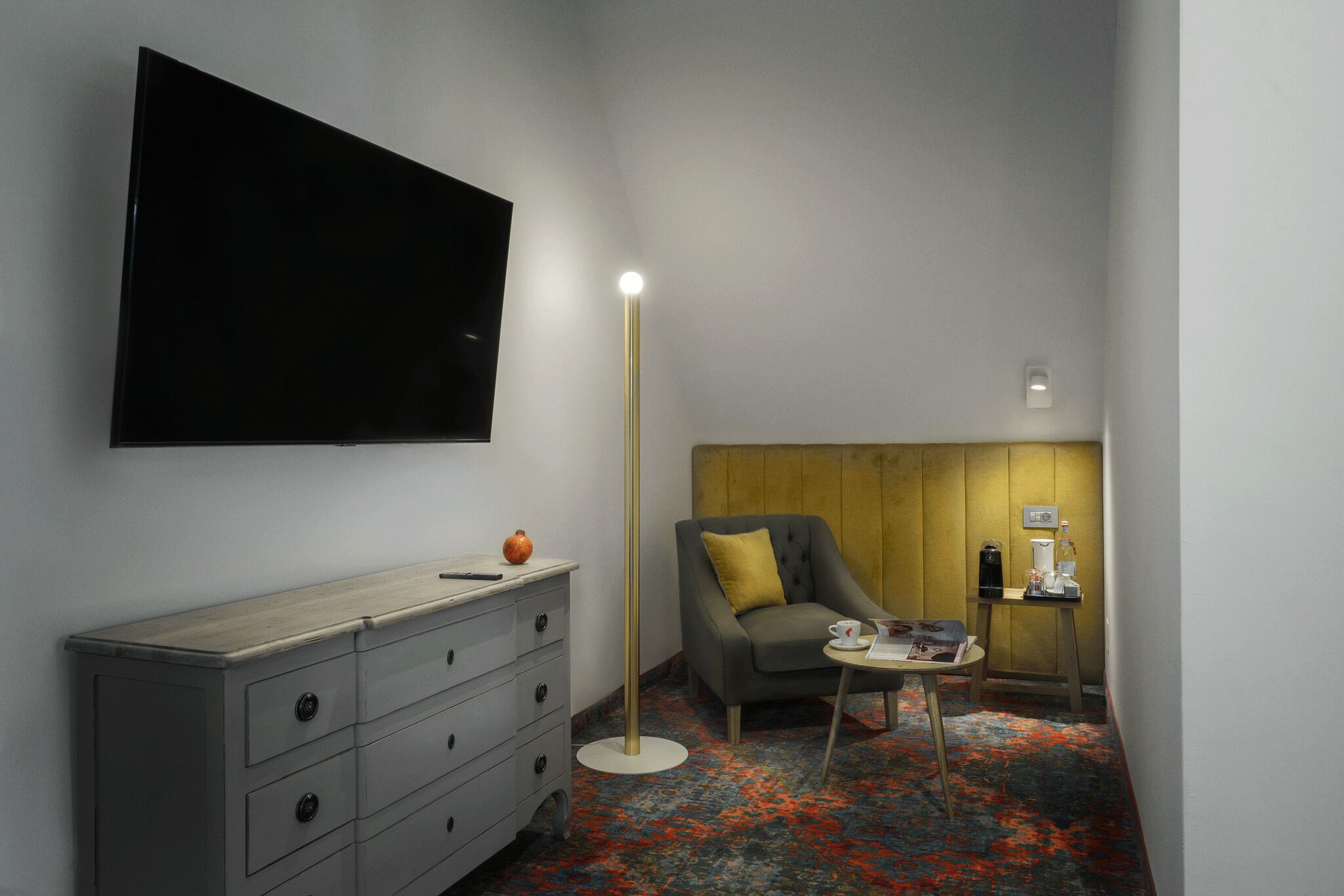 A hotel room with a flat-screen TV mounted on the wall, a grey dresser with drawers, a grey armchair with a yellow pillow, a small round table with a lamp, and a coffee machine on a wooden stand.