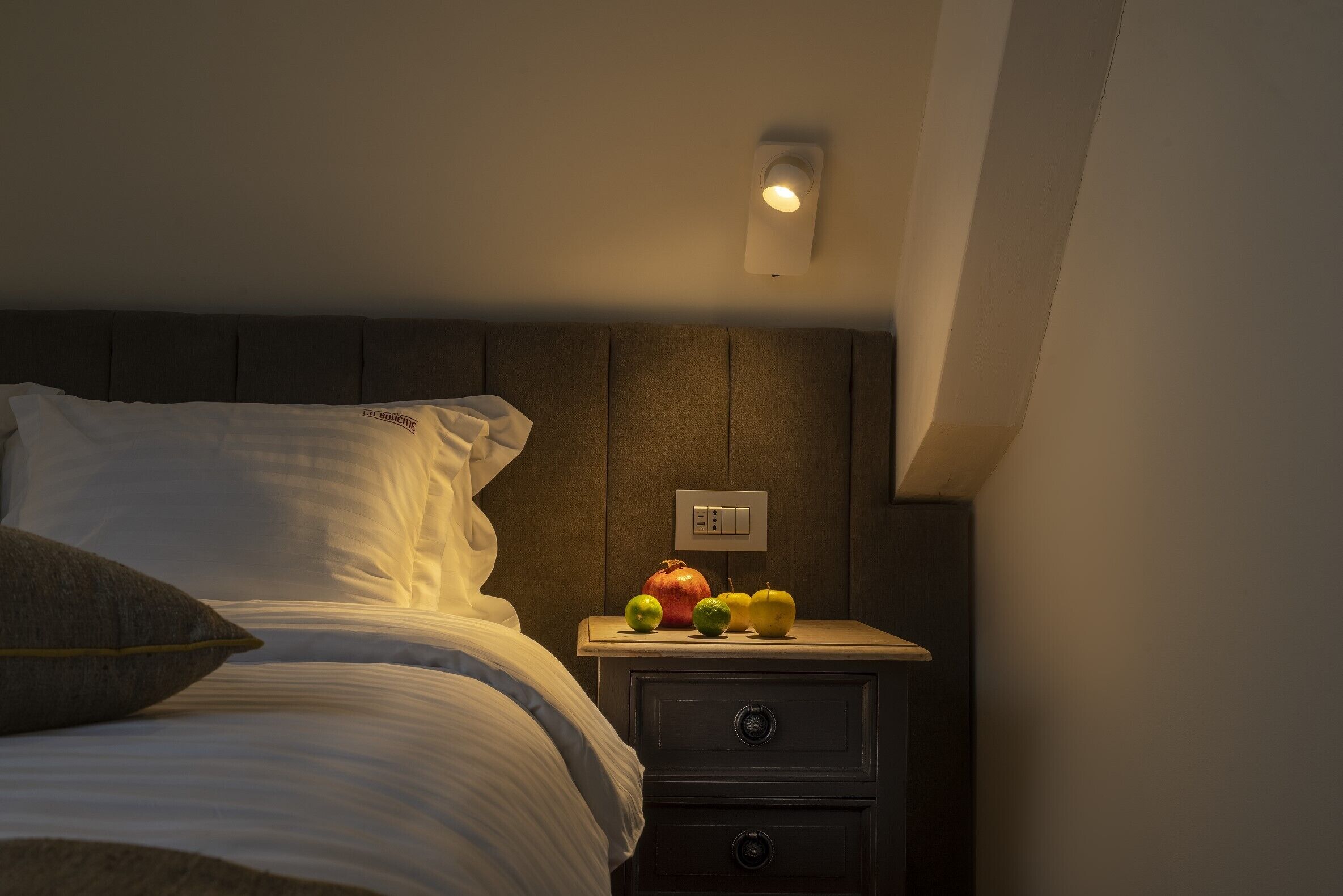 A neatly made bed with white linens, a dark headboard, and a bedside table with a fruit bowl containing apples and a pomegranate.
