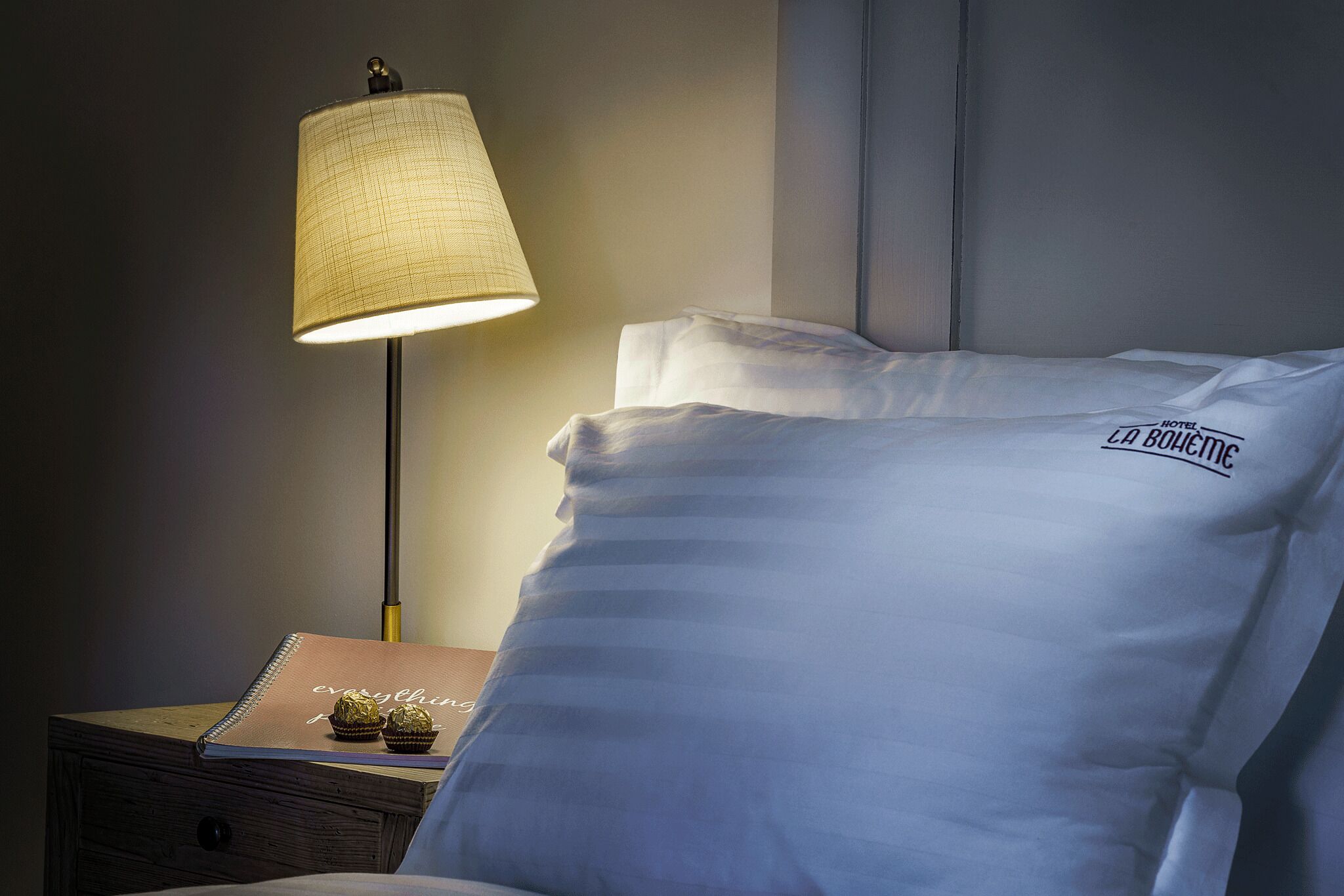 A neatly made bed with a pillow labeled "Hotel La Bouviere," a bedside lamp, and a small table with a notebook and chocolates.